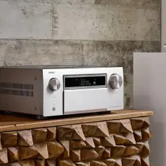 Srebrny amplituner Denon z wyświetlaczem Dolby Atmos, obok białej kolumny głośnikowej. To centrum domowego kina, które pozwala cieszyć się kinową jakością dźwięku.