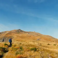 Escursionista in cammino verso il monte più alto dell'Emilia Romagna, con un paesaggio dorato e un cielo azzurro.