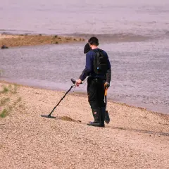 Mężczyzna z wykrywaczem metali i łopatką na plaży przy wodzie.