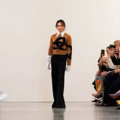 Victoria Beckham går på catwalken, klädd i en brun tröja med ett svart mönster och vida svarta byxor. Publiken, inklusive Harper Beckham, fotograferar med sina mobiler.