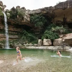 Niños disfrutan en una **piscina natural** bajo una cascada, rodeada de rocas y vegetación.