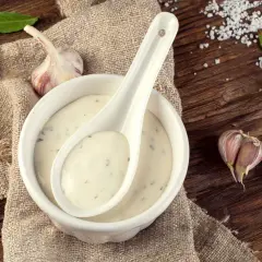 Sauce à l'ail crémeuse dans un bol blanc, avec une cuillère, des gousses d'ail et des feuilles de laurier sur une table en bois rustique.