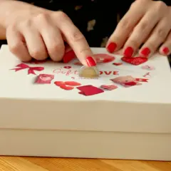 Dłonie z czerwonymi paznokciami wkładają monetę do pudełka z napisem "Gift LOVE".