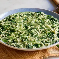 Un cremoso risotto con ortiche, servito in un piatto blu, con forchetta e coltello a lato. Un pezzo di parmigiano e una grattugia completano la scena.