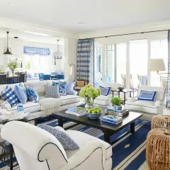 A bright living room with coastal interior design, featuring white sofas, blue and white patterned pillows, and a dark wood coffee table.