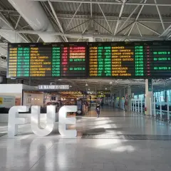 Tablica odlotów na lotnisku, z widocznym napisem "FUE". Informacje o lotach, w tym boarding, wskazują na podróż.
