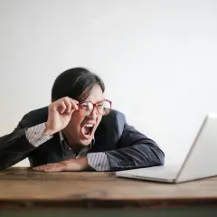 Mann mit Brille schreit frustriert vor Laptop. Vielleicht lernt er gerade, wie funktioniert ein Fernstudium, und ist überfordert.