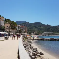 Promenad längs stranden i Port de Sóller med folk som njuter av solen och havet, omgivet av gröna berg.