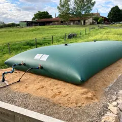 Gran depósito de agua de 20 litros, verde, instalado en un campo con una granja al fondo.