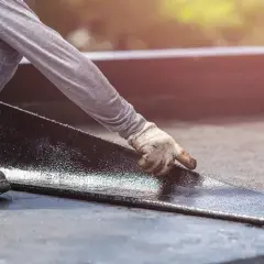 Hand mit Handschuh legt neue Dachpappe an. Alte Dachpappe entsorgen ist wichtig für die Langlebigkeit.