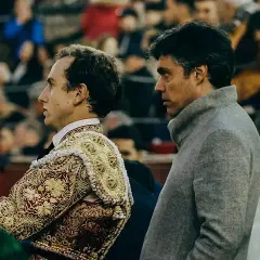 Un torero con traje de luces bordado y zapatillas torero, junto a un hombre con abrigo gris, observan la plaza.