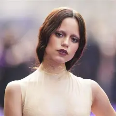 Jenna Ortega, czy ma polskie korzenie? Aktorka w kremowej sukni z siateczkowym kołnierzem, z burzą brązowych włosów i wyrazistym makijażem.