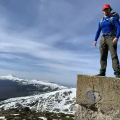 Hombre con mochila en la cima de un pico nevero, con vistas a montañas nevadas bajo un cielo azul con nubes.