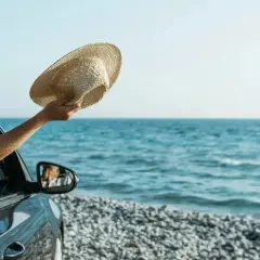 Ręka z kapeluszem wychyla się z samochodu na tle morza. Idealne wakacje leasingowe!