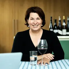Eine lächelnde Frau, die wie ein **master of wine** wirkt, sitzt an einem Tisch mit Wein und Wasser. Im Hintergrund stehen mehrere Weinflaschen.