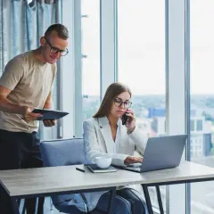Un homme consulte un document pendant qu'une femme au téléphone travaille sur son ordinateur portable, illustrant l'efficacité de l'e-commerce B2B.