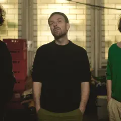 Trzech mężczyzn, w tym Andrzej Konieczny, pozuje do zdjęcia. Jeden z nich ma kręcone włosy i wąsy, drugi brodę, a trzeci długie włosy. W tle widać ceglaną ścianę i jakieś pudła.