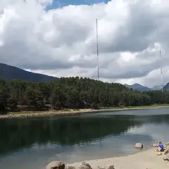 Persona sentada en la orilla del lago de Engolasters, rodeado de pinos y montañas, con dos torres de telecomunicaciones al fondo.