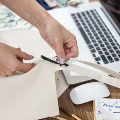 Ręce szyją torbę shopper, dodając czarną wstążkę. Na biurku laptop, myszka i materiały do szycia.