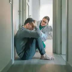 Lekarz wspiera pacjenta zmagającego się ze splątaniem w psychiatrii. Mężczyzna siedzi na podłodze, przygnębiony, a lekarka pochyla się nad nim z troską.