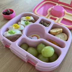 Różowa śniadaniówka do szkoły z przegródkami zawiera zawinięte tortille z uroczymi ozdobami, winogrona, orzeszki pistacjowe, krakersy i suszone mango.