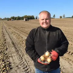 Rolnik trzyma świeżo zebrane cebule na polu. W tle widać rzędy wykopanych warzyw, maszyny rolnicze i drzewa. To prawdziwe klasy ziemi.