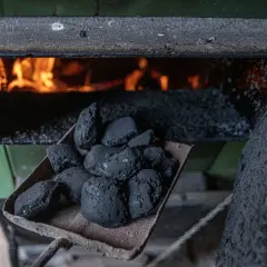Łopata z węglem przed rozpalonym piecem bezklasowym. Płomienie żarzą się w palenisku, a obok leży bryła węgla.