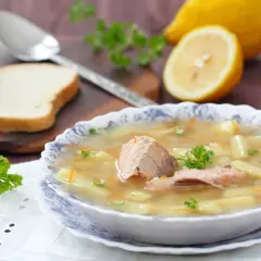 Miseczka zupy śledziowej z ziemniakami i marchewką, ozdobiona natką pietruszki. Obok cytryna i kromka chleba.