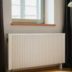 Vitt element med ribbad front, perfekt för att bygga elementskydd. Bredvid fönster med mörka gardiner och en golvlampa.