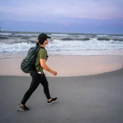 Kobieta z plecakiem na plaży, ćwicząca rucking przy falach oceanu.