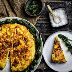 Eine goldbraune Tortilla de Patatas, umgeben von gebratenen Pimientos de Padrón. Daneben eine Portion Salz und ein Stück auf einem Teller.