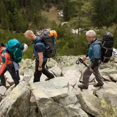 Drei Wanderer mit Rucksäcken und Helmen auf felsigem Untergrund. Die **Packliste Klettersteig** ist gut gefüllt für das Abenteuer.