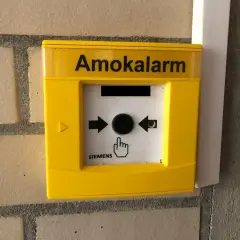 Gelber Amokalarm-Knopf an einer Ziegelwand. Ein Symbol zeigt eine Hand, die auf den Knopf drückt, um einen Amoklauf an einer Schule zu melden.