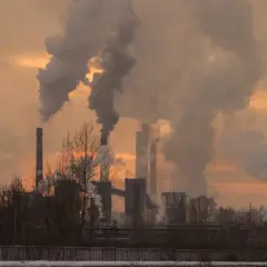 Chimeneas industriales expulsan humo denso, creando un paisaje sombrío. Las partículas contaminantes se elevan, tiñendo el cielo de un naranja opaco.