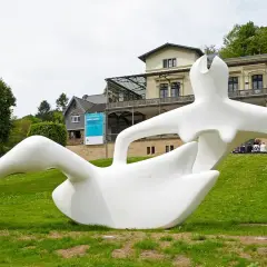 Weiße Skulptur einer liegenden Figur im Freien vor einem Gebäude mit der Aufschrift "Bahnhof Rolandseck".