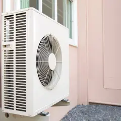 Unité extérieure d'une pompe à chaleur, installée sur un mur rose. Le ventilateur est visible, prêt à assurer le confort thermique.