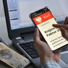 Dłoń trzyma smartfon z ekranem KSeF, obok leżą pieniądze i kalkulator.