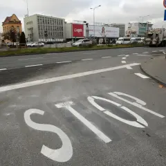 Na asfalcie widoczna jest biała linia warunkowego zatrzymania "STOP". W tle budynki i pojazdy.