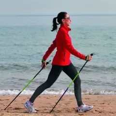 Kobieta w czerwonej kurtce ćwiczy marsz z kijkami na plaży. Pokazuje, jak chodzić z kijkami, wykorzystując je do aktywności fizycznej nad morzem.