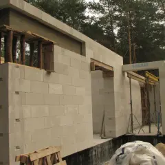 Budowa domu z bloczków betonowych. Widoczna ściana nośna z otworami okiennymi i drzwiami, podparta drewnianymi belkami.