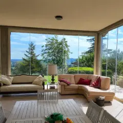 Soggiorno luminoso con divani in pelle e tessuto, tavolo in legno e ampie vetrate che incorniciano un panorama verde. Idee case con vetrate per godersi la natura.