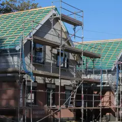Baugerüste umgeben Häuser im Rohbau, die Dächer mit grüner Unterspannbahn und Holzlatten für die Ziegel zeigen. Die Satteldach Konstruktion ist gut erkennbar.