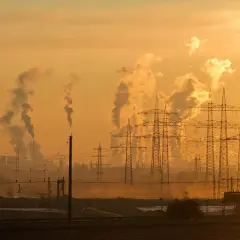 Dymiące kominy fabryk i linie energetyczne na tle pomarańczowego, zachodzącego słońca.