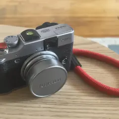 Srebrny aparat Fujifilm X20 z czerwoną smyczą leży na drewnianym blacie.
