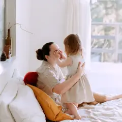Dziecko dotyka nosa mamy, uosabiając wychowanie bliskościowe. Czuły moment na łóżku, otoczonym jasnymi poduszkami i światłem.