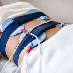 Osoba leży pod kocem z pasami i elektrodami na brzuchu, przygotowana do zabiegu ems trening.