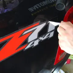 Mano che rimuove adesivo "Z7 4x4" da auto nera, usando un phon per ammorbidire il nastro adesivo vecchio.