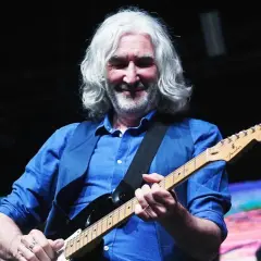 Muzyk z siwymi włosami gra na gitarze elektrycznej, w niebieskiej koszuli i kamizelce. W tle widać ekran z kolorowym obrazem, przypominającym krajobraz, jakby z czasów Dire Straits.