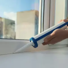 Ręce trzymają pistolet z uszczelniaczem, aplikując go na biały parapet okna.