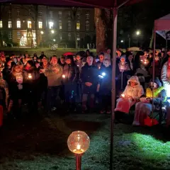 Menschen versammeln sich bei Kerzenlicht zu einem Gottesdienst am Heiligabend vor einem Gebäude. Ein rotes Kreuz steht im Vordergrund.